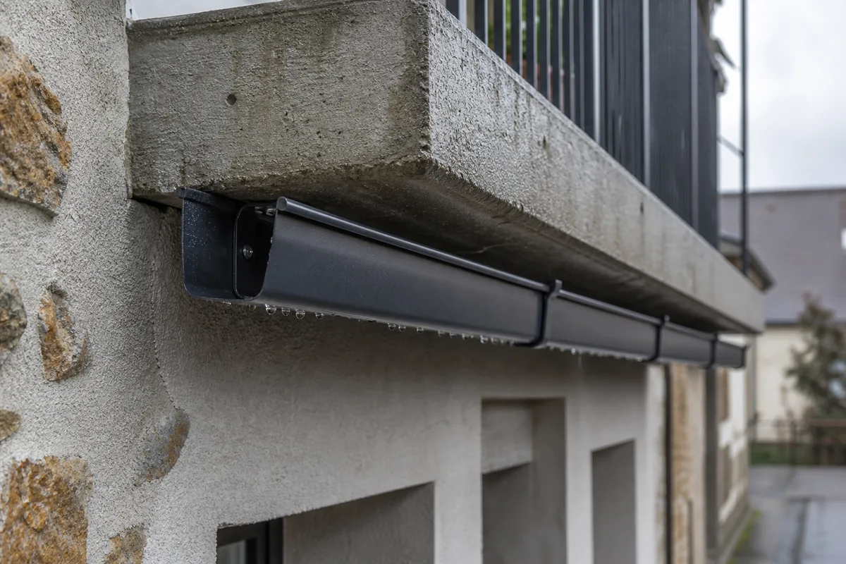 Gouttière aluminium gris anthracite fixée sous le nez de dalle d’un balcon, façade en moellon enduit sous une lumière voilée