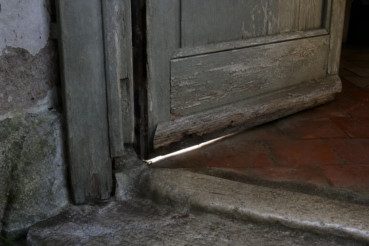 Ancienne porte d’entrée en bois plein à la peinture écaillée, jour visible sous le seuil de pierre usé